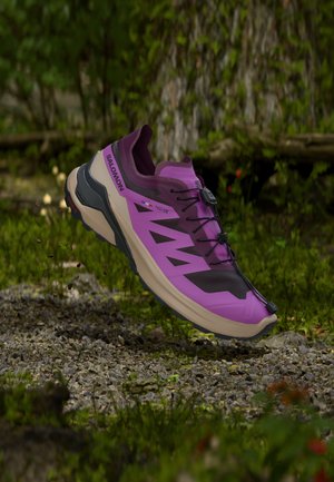 Purple and black Salomon trail running shoe airborne above a gravel path with forest greenery in the background.