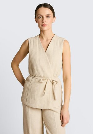 Kobieta w beżowym bezrękawnym topie wiązanym w talii i pasujących szerokich spodniach na jednolitym tle.