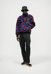 Colorful fleece pullover with a blue, red, and black pattern, featuring a zip neck and a black chest pocket. Paired with black pants and sneakers.