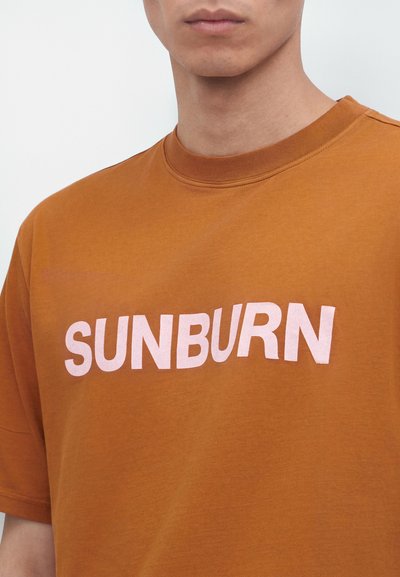 Άνδρας φοράει καφέ μπλουζάκι με τη λέξη «SUNBURN» τυπωμένη με μεγάλα λευκά γράμματα στο στήθος.