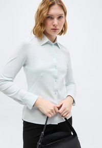 Γυναίκα με κοντά ξανθά μαλλιά, φορώντας ανοιχτό μπλε πουκάμισο με κουμπιά και κρατώντας μια μαύρη τσάντα, σε απλό φωτεινό φόντο.