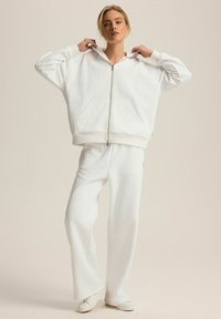 Biała, oversize'owa bluza z kapturem na zamek, z przednimi kieszeniami, w zestawie z luźnymi białymi spodniami. Gładka struktura materiału. Prosty design, bez wzorów.