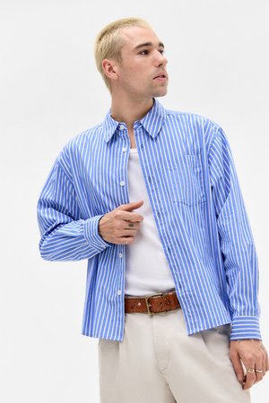Un homme aux cheveux blonds courts porte une chemise rayée bleue et blanche sur un débardeur blanc, un pantalon beige et une ceinture marron, regardant sur le côté.