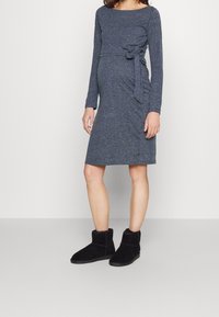 Tmavě šedé těhotenské šaty s dlouhým rukávem, bočním vázáním, délka po kolena a jemná textura, doplněné černými kotníkovými botami.