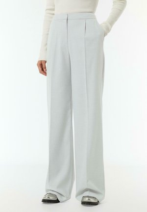 Pantalons large gris clair avec plis avant, fabriqués dans un tissu lisse. Dotés de poches latérales et d'une taille haute. Portés avec des chaussures argentées.