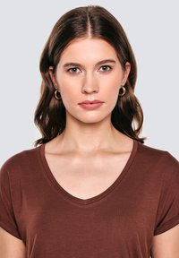 Brązowy t-shirt z dekoltem w serek i krótkimi rękawami, wykonany z miękkiej, teksturowanej tkaniny. Modelka ma średniej długości falowane włosy i nosi złote kolczyki koła.