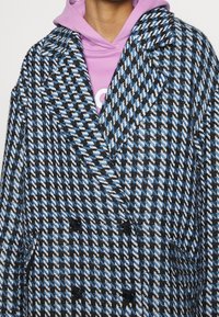 Dvojité sako s modro-černobílým houndstooth vzorem. Vyrobeno z texturované látky s černými knoflíky a detaily límce.