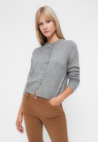 Siv cardigan iz volne z tremi srebrnimi zategovalnimi zapiralkami spredaj. Ima okroglo izrez in rebraste manšete, kombiniran z rjavimi žamtnimi hlačami.