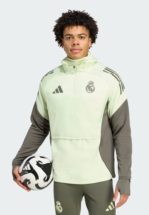 Light green athletic jacket with dark gray accents, zippered collar, and team logo. Model holds a black and white soccer ball.