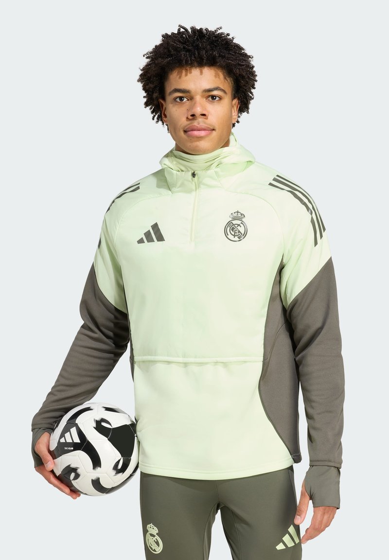 Hellgrüne Sportjacke mit dunkelgrauen Akzenten, mit Reißverschlusskragen und Teamlogo. Das Modell hält einen schwarz-weißen Fußball.