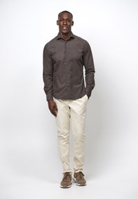 Camicia marrone con bottoni, pantaloni crema e sneakers marroni. La camicia ha una texture liscia; l'outfit presenta una vestibilità casual senza motivi.