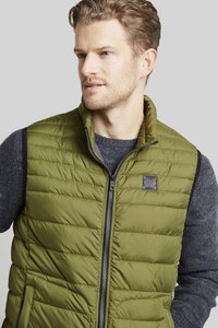 Olivgrüne isolierte Weste mit gestepptem Muster, Reißverschluss vorne und schwarzen seitlichen Akzenten. Verfügt über ein Logopatch auf der Brust.