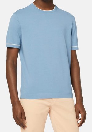 Mann trägt hellblaues kurzärmeliges T-Shirt mit weißem Rand an Ärmeln und Hals, kombiniert mit beigen Hosen, stehend vor weißem Hintergrund.