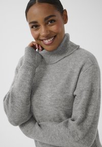Pull à col roulé gris en matériau doux et texturé, avec des poignets côtelés et une coupe décontractée, mettant en avant un design à la fois confortable et moderne.