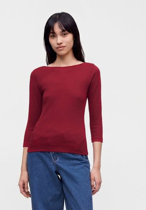 Junge Frau mit glatten schwarzen Haaren, die ein eng anliegendes dunkelrotes Langarmshirt und blaue Jeans trägt, steht vor einem schlichten hellen Hintergrund.