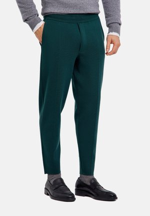 Hombre con pantalones oscuros verdes a medida, calcetines grises, mocasines negros de piel, jersey gris y camisa blanca con las manos parcialmente en los bolsillos.