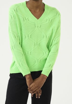 Persona vistiendo un suéter de cuello en V texturizado de color verde claro con mangas largas, de pie con las manos entrelazadas al frente, combinado con pantalones negros.