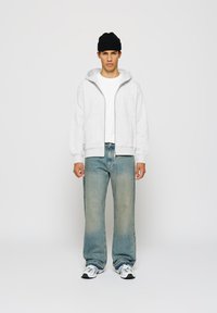Ung mann som står, iført en lys grå zip-up hettegenser, hvit t-skjorte, blå jeans, hvite joggesko og en svart beanie mot en enkel bakgrunn.