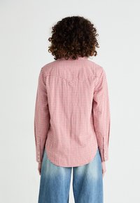 Chemise à manches longues rouge et blanche à carreaux avec des poignets à boutons, présentant un ourlet arrondi et un détail de empiècement au dos. Associée à un jean bleu ample.