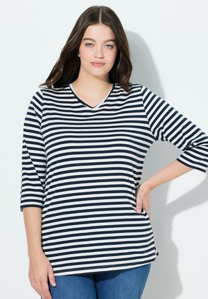 Mladá žena s dlouhými hnědými vlasy, oblečená v černobílém pruhovaném topu a modrých džínách, stojící s jednou rukou na boku na bílém pozadí.