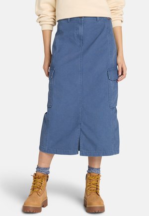 Jupe midi en coton bleu avec fente avant et poches latérales, associée à des bottes à lacets beiges et des chaussettes à motifs. Design simple et utilitaire.
