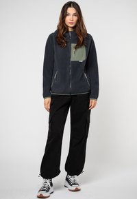 Fleece zip-up jack in donker marineblauw met een licht olijfgroen borstzakje. Voorzien van ritszakken aan de zijkanten en contrasterende groene afwerking.