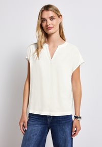 Weiße kurzärmelige Bluse mit V-Ausschnitt und lockerer Passform, aus glattem Stoff, kombiniert mit blauen Jeans und einem silbernen Armreif.