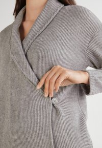 Grijze geribbelde cardigan met een sjawlkragen, voorzien van een enkele knoopsluiting. Zachte textuur en ontspannen pasvorm, die de warmte van de stof benadrukt.