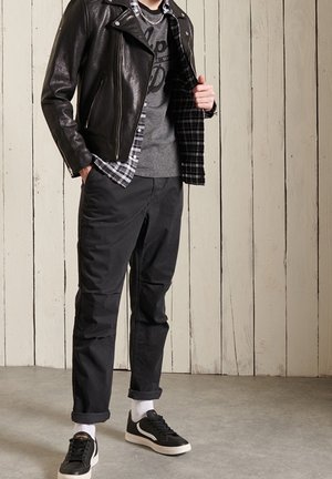 Hombre vistiendo una chaqueta de cuero negra sobre una camisa a cuadros y una camiseta gris, pantalones negros arremangados en los puños, zapatillas negras, de pie en interiores.