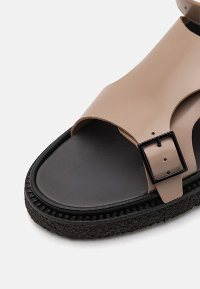 Beige faux leather sandal with an open toe, adjustable black buckle, thick black textured sole, and smooth black footbed.