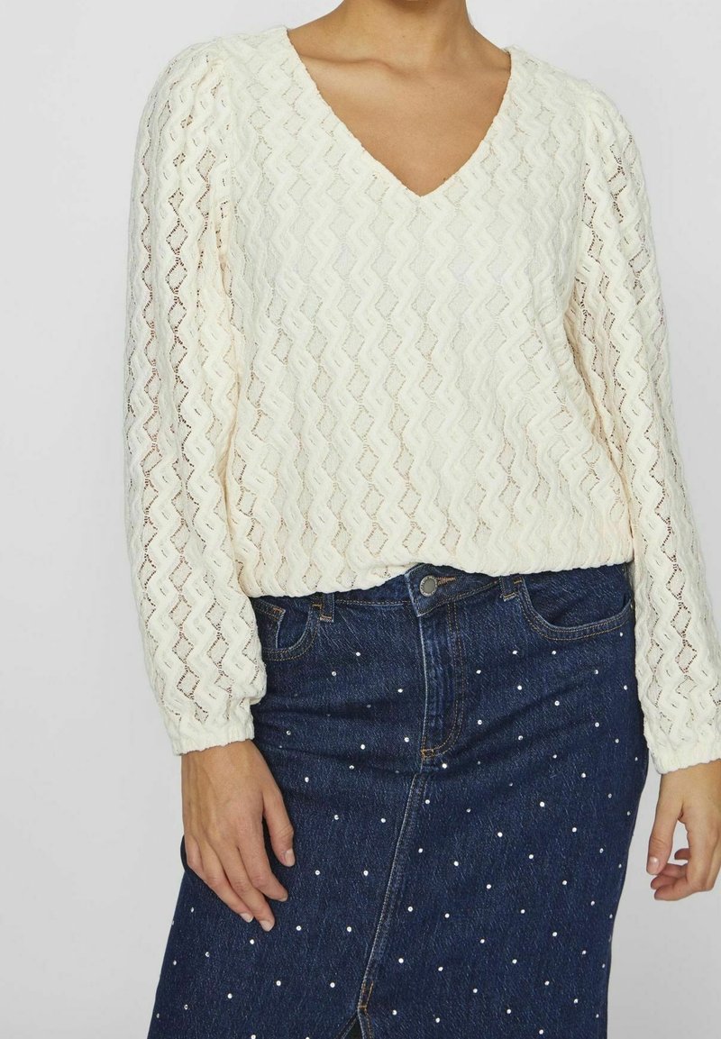 Femme portant un chemisier blanc texturé à manches longues avec un col en V et une jupe en denim bleu foncé décorée de petits points blancs.