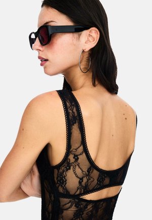 Schwarzes Spitzen-Bodysuit mit transparenter Textur, gewelltem Rand und offenem Rückendesign; kombiniert mit übergroßen schwarzen Sonnenbrillen und Ohrhängern.
