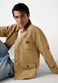 Jonge man met donker haar, gekleed in een tan kleurige jas met voorzakken en een wit shirt, zittend tegen een eenvoudige lichte achtergrond.