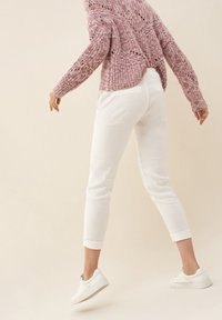 Rosa gestrickter Pullover mit lockerer Passform und Lochmuster, kombiniert mit weißen, schmal zulaufenden Hosen und weißen Sneakers, die einen lässigen Stil zeigen.