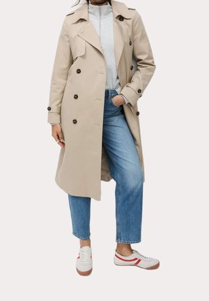 Persona que lleva un abrigo trench beige de doble botonadura, parte superior gris clara con cremallera, vaqueros azules y zapatillas blancas con rayas rojas, de pie con una mano en el bolsillo.