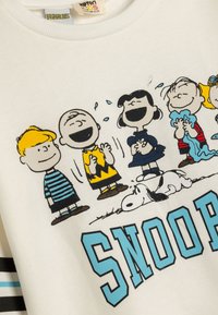 Biała bluza z kapturem z postaciami z komiksu Peanuts, z czarnymi konturami i kolorowymi akcentami. "SNOOPY" nadrukowane dużymi niebieskimi literami.