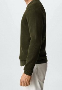 Pull tricoté vert olive avec texture côtelée, encolure en V et poignets ajustés. Porté avec un pantalon de couleur claire, tenu par une silhouette masculine.