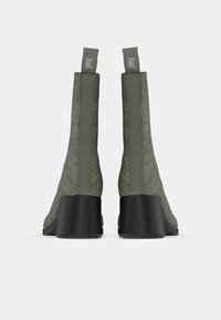 Par de botas de tobillo de tela verde oliva con tacones de bloque negros vistas desde la parte posterior sobre un fondo blanco.