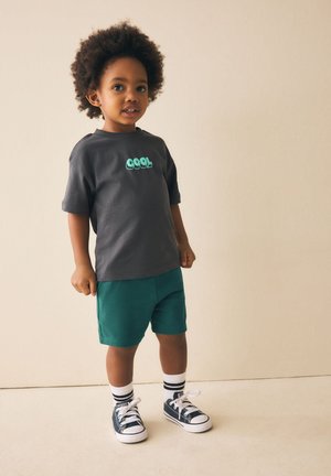 Nuori lapsi kiharatukka, joka käyttää tummaa "COOL"-t-paitaa, vihreitä shortseja, raidallisia sukkia ja mustia tennareita, seisoo yksiväristä taustaa vasten.