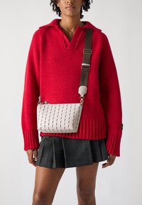 Svetlosivý crossbody batoh s textúrovaným vzorom a geometrickými výrezmi. Nastaviteľný hnedý popruh, kombinovaný s červeným pleteným sveterom a kockovanou sukňou.