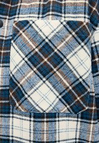 Chemise en flanelle avec un motif à carreaux bleu, marron et blanc ; comprend une poche poitrine avec un design assorti et une texture douce.