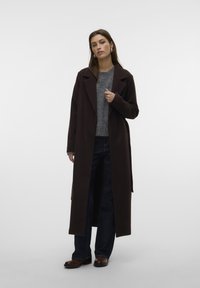 Cappotto lungo marrone con una texture liscia, caratterizzato da un colletto a punte e apertura frontale, indossato sopra un maglione grigio a maglia e jeans scuri.