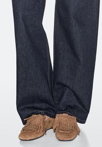 Brown suede moccasin-style shoes with fringe accents, worn with dark blue denim pants showcasing a straight leg cut and visible stitching.