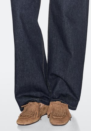 Brune mokkasinstilssko i ruskind med frynseaccenter, båret med mørkeblå denimbukser med rette ben og synlige syninger.