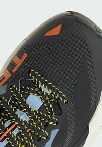 Chaussure de sport noire avec une tige texturée, des lacets colorés avec des accents jaunes, des motifs géométriques et un détail orange sur le côté.