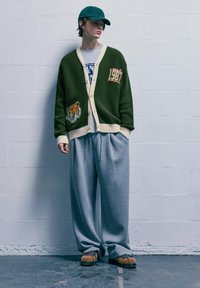 Cardigan verde con rifiniture e toppe crema, pantaloni felpati grigi oversize, accentuati da scarpe marroni texture e un cappellino verde.