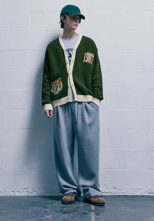 Zöld kardigán krém szegéllyel és foltokkal, szürke oversized melegítőnadrág, megerősítve textúrával rendelkező barna cipővel és zöld sapkával.