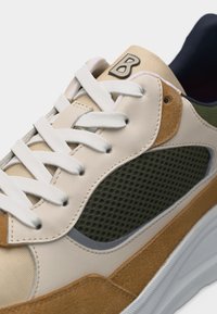 Κοντινή λήψη του μπεζ, καφέ και πράσινου αθλητικού παπουτσιού με λευκά κορδόνια, δίχτυ στο πλάι, σουέτ λεπτομέρειες και λογότυπο "B" στη γλώσσα.