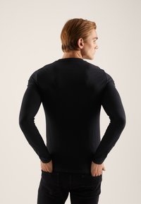 Črna majica z dolgimi rokavi iz gladkega materiala, oprijet kroj, z okroglim izrezom in brezšivnimi rameni, nošena s temnimi hlačami.