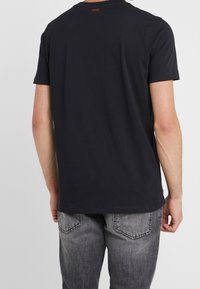 Camiseta de algodón negra de manga corta, con un escote redondo y un logotipo rojo en la parte posterior. Combinada con vaqueros de mezclilla grises.